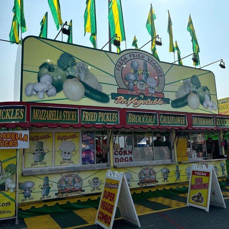 Dr. Vegetable food stand at a fair with green and yellow flags, serving fried vegetables, pickles, and corn dogs.