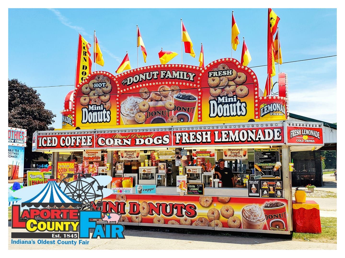 A colorful carnival food stand at the LaPorte County Fair selling mini donuts, iced coffee, corn dogs, and lemonade.