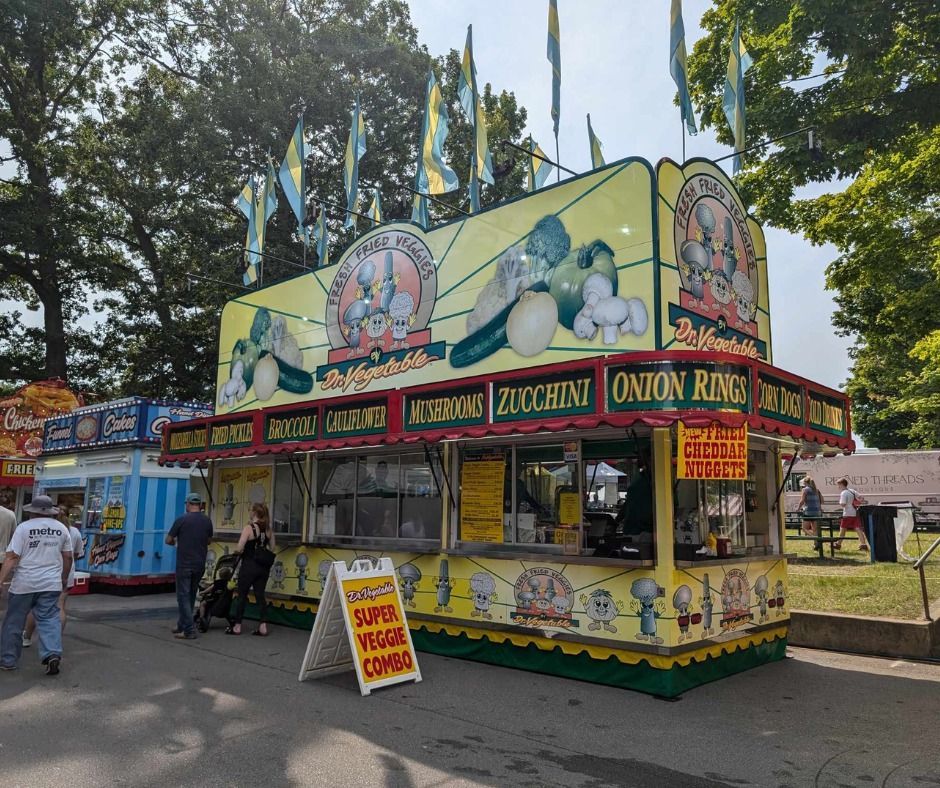 A yellow food stand at a fair featuring painted vegetables, signs for fried foods, and a 