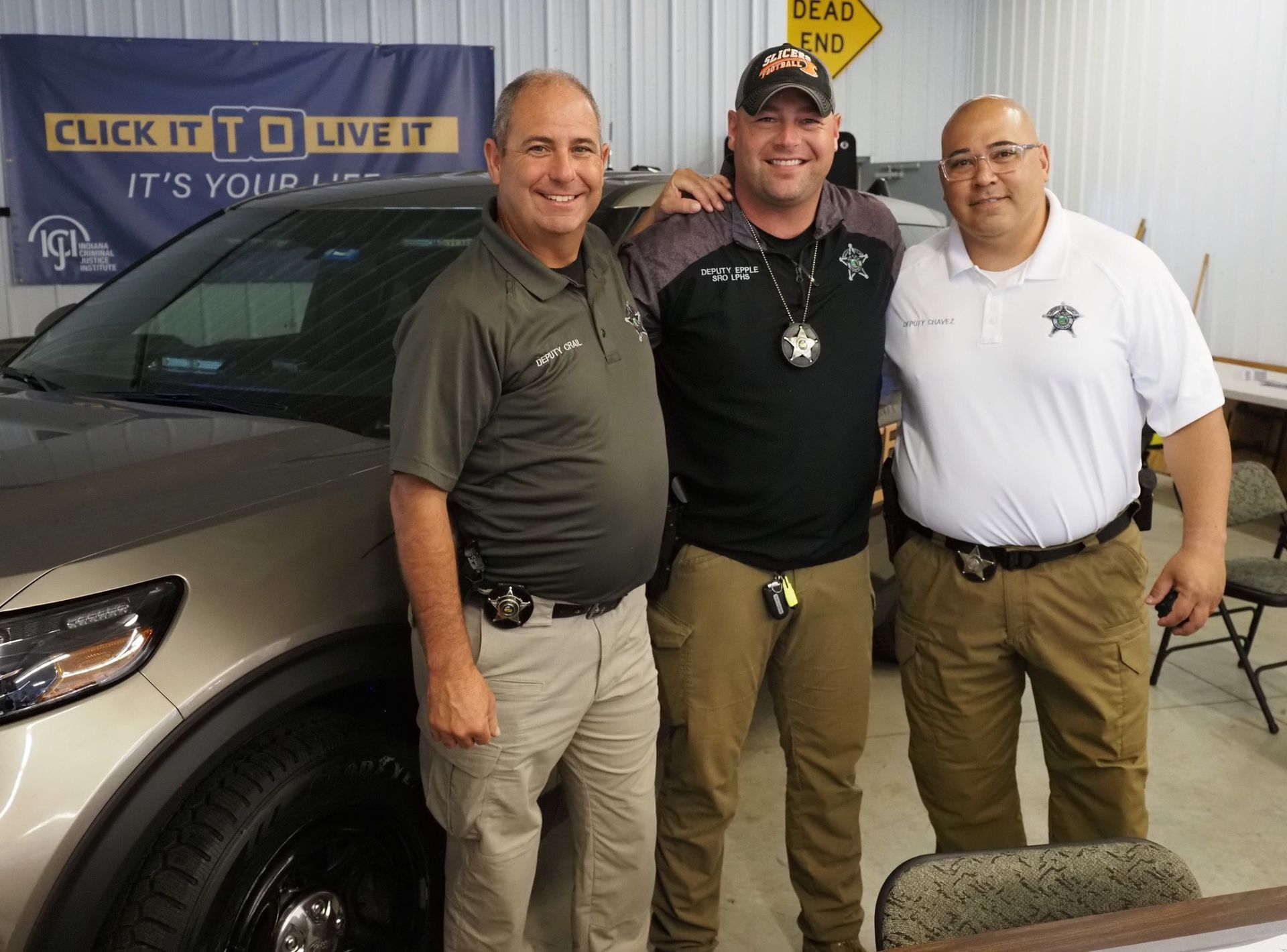 Three law enforcement officers stand smiling in a room next to a police vehicle and a