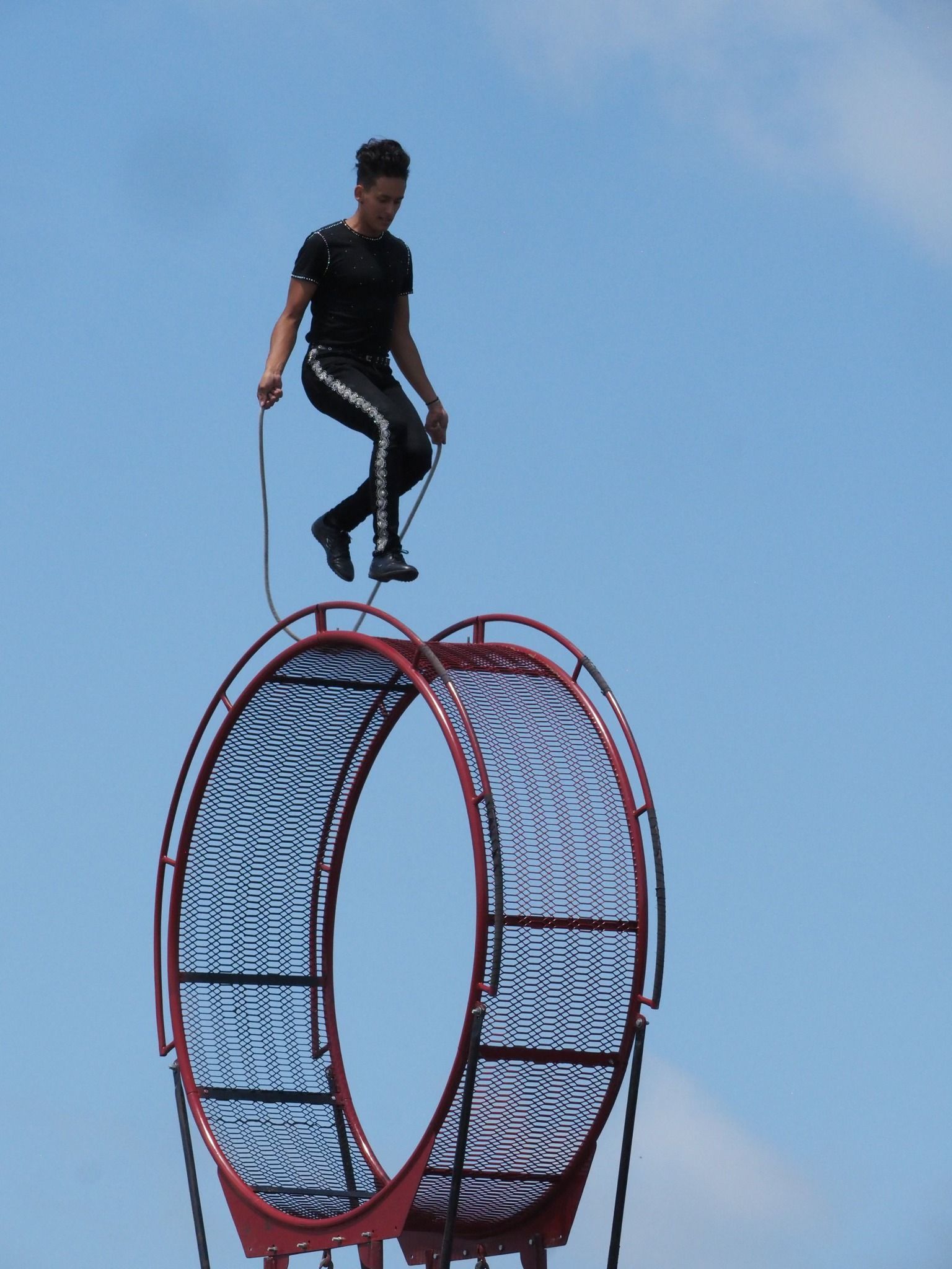 A person in dark athletic clothing jumps rope on top of a red, circular metal apparatus against a blue sky.