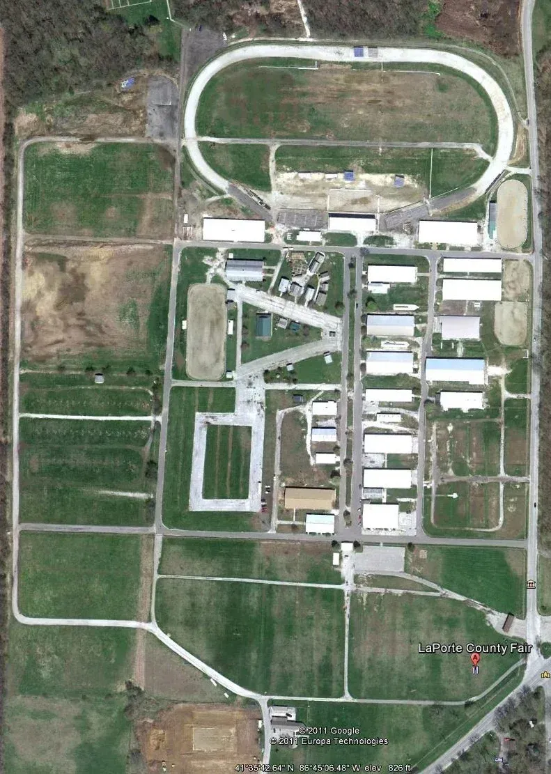 Aerial view of an agricultural fairgrounds complex featuring a large racetrack, barns, and open grassy fields.