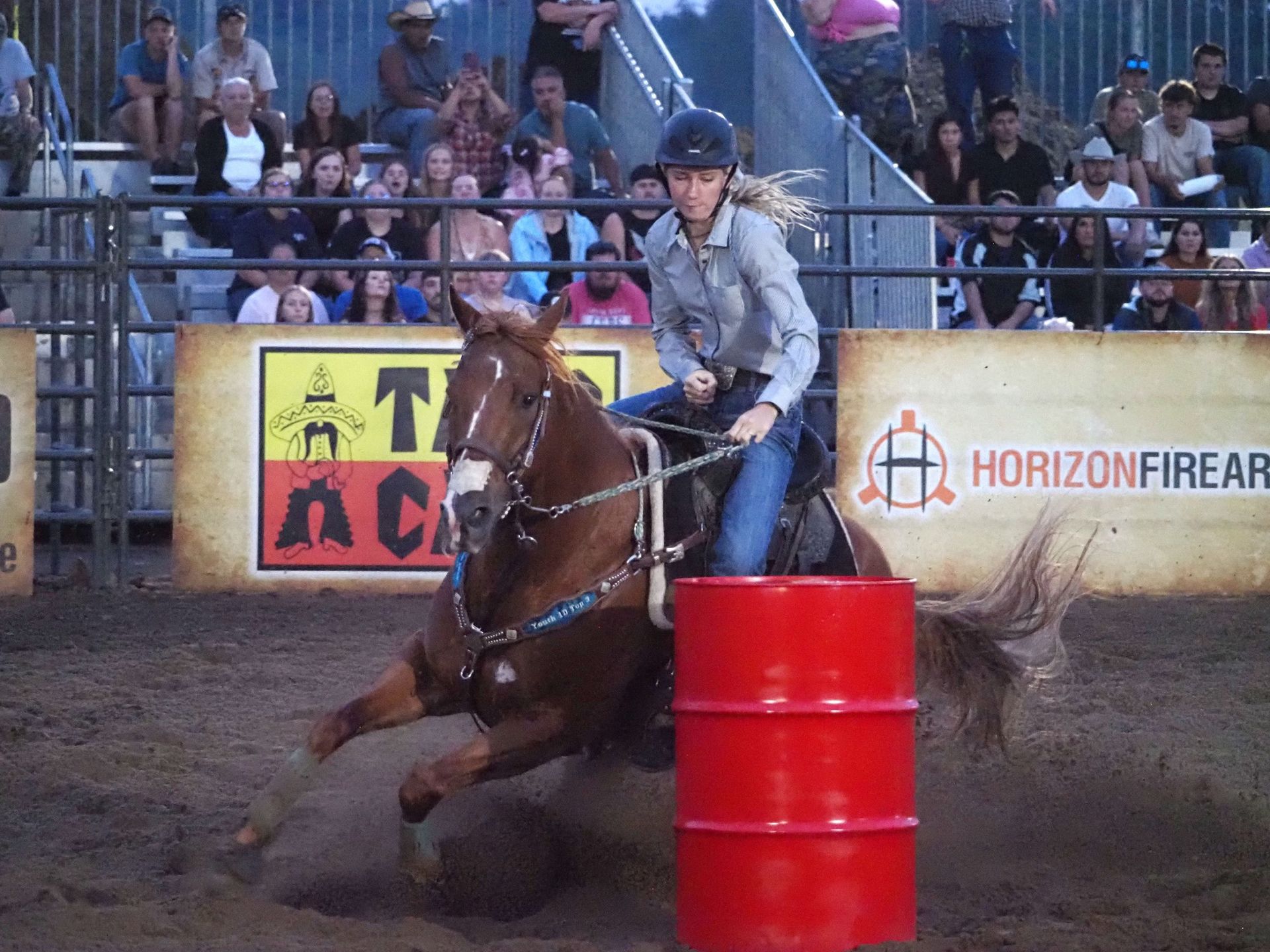 A rider in a helmet steers a brown horse around a bright red barrel during a competition in a dirt arena.