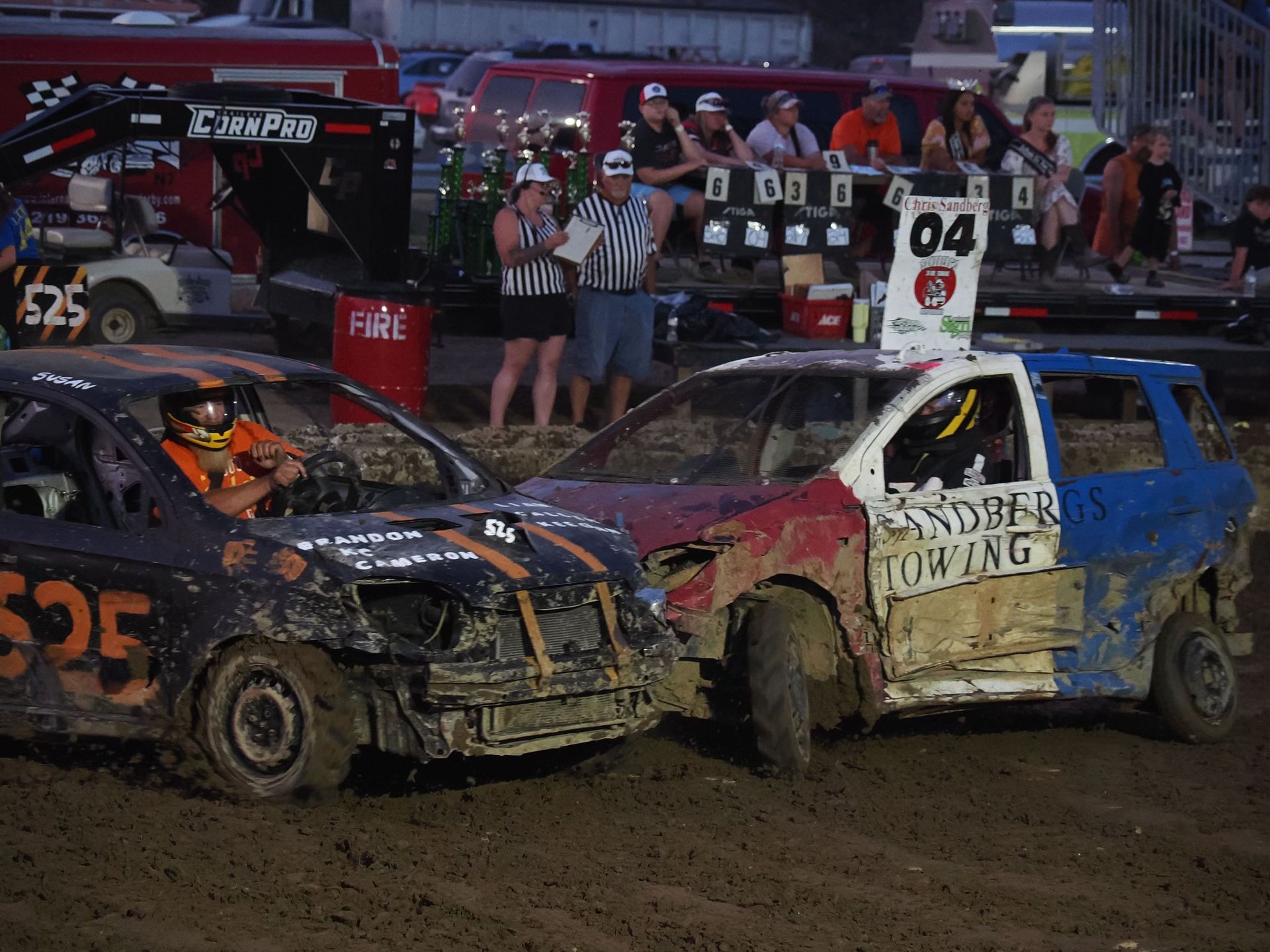Two cars collide in a dirt track demolition derby, with spectators watching from the background.