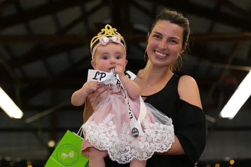 A smiling person holds a baby wearing a decorative crown and a pink dress with a sash in an indoor setting.