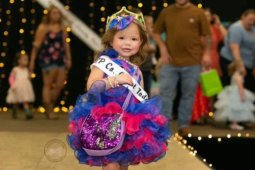 A child wearing a crown and a blue and pink ruffled dress with a sash, holding a sequined purse on a stage with lights.