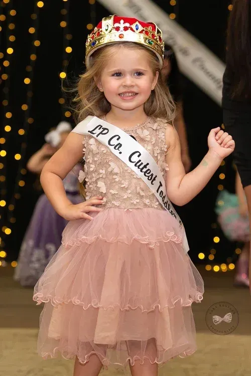 A child wearing a crown and a dress with a sash that reads, 
