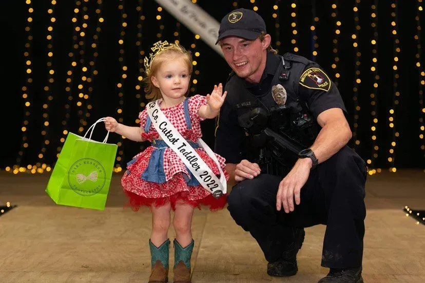 A toddler in a pageant sash and cowgirl boots waves while standing next to a uniformed officer against a lit backdrop.