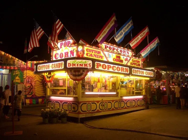 A brightly lit concession stand at a night fair serving popcorn, cotton candy, and drinks, decorated with flags.