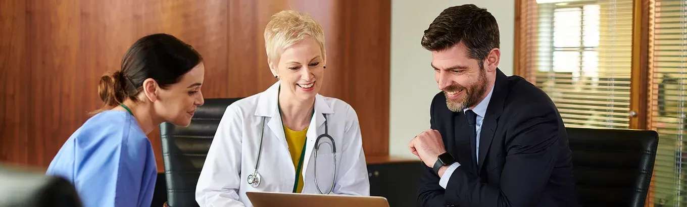 Three people, a nurse, doctor, and businessman, review a laptop. The doctor wears a stethoscope, and the businessman smiles.