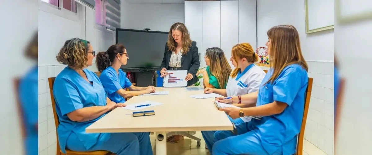 A group of nurses in blue scrubs at a meeting with a woman presenting charts, inside a room.