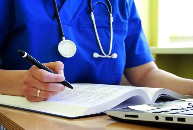 Person in blue scrubs writing in a notebook, stethoscope around neck, laptop visible on desk.