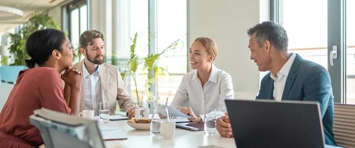 Three people in business attire at a table in a meeting. A woman is speaking with two others.