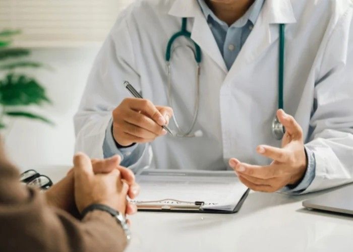 A doctor in a white coat with a stethoscope around their neck sits at a desk, gesturing while talking to a patient.