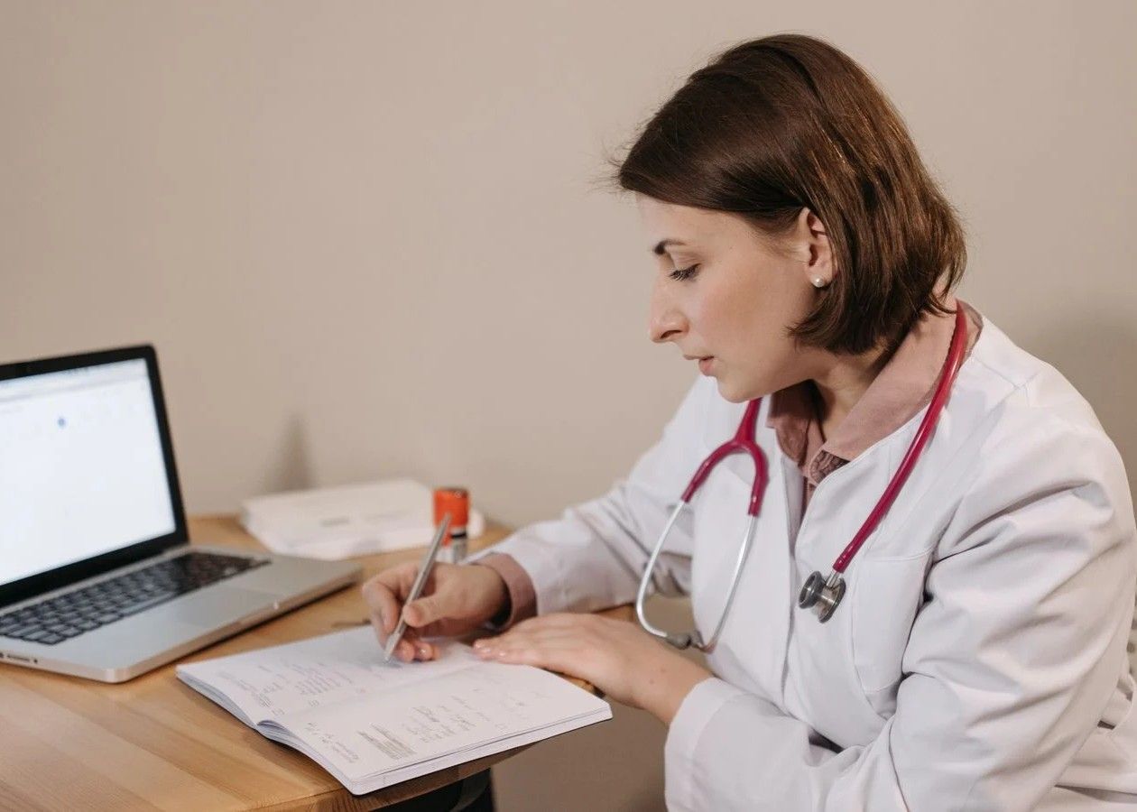 Doctor in a white coat with a stethoscope writes notes at a desk beside a laptop.