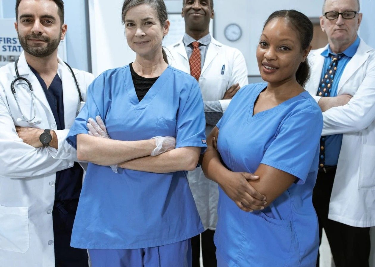 Group of medical staff in blue scrubs and white coats standing together in a hospital hallway, arms crossed.