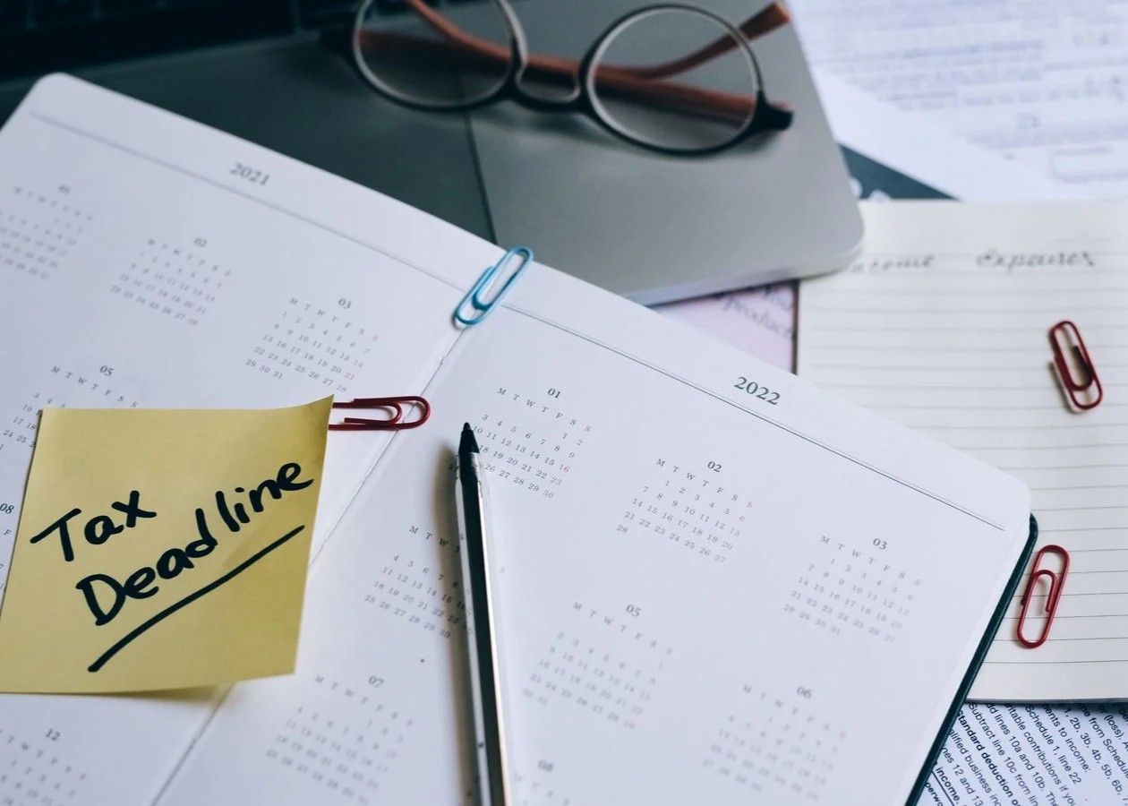 Desk with open planner, paper clips, glasses, and yellow sticky note reading “Tax Deadline.”