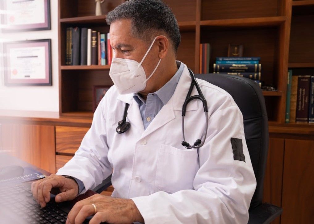 Masked doctor in white coat using a laptop at a desk in an office