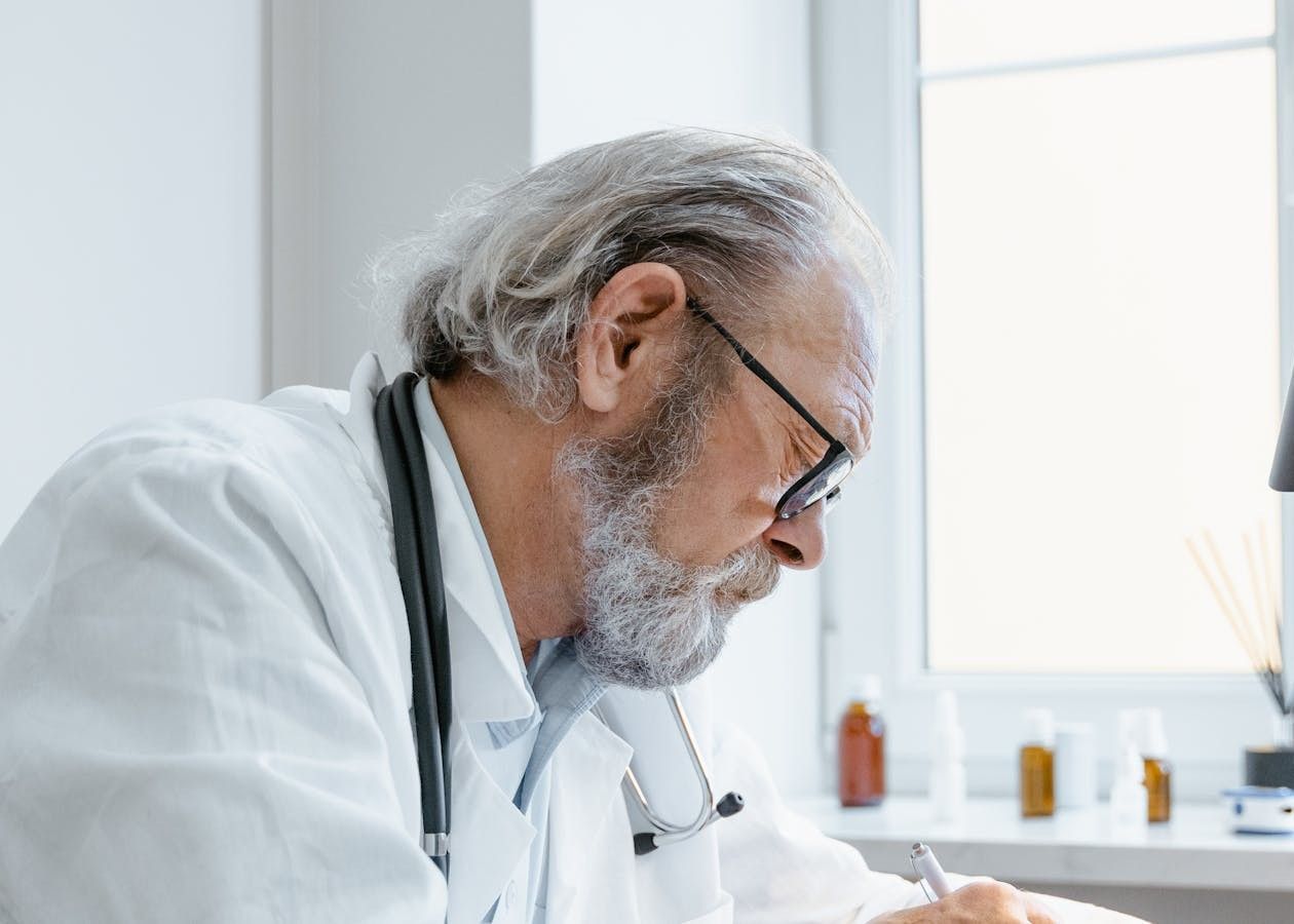 Doctor in a white coat writing notes at a desk beside a bright window