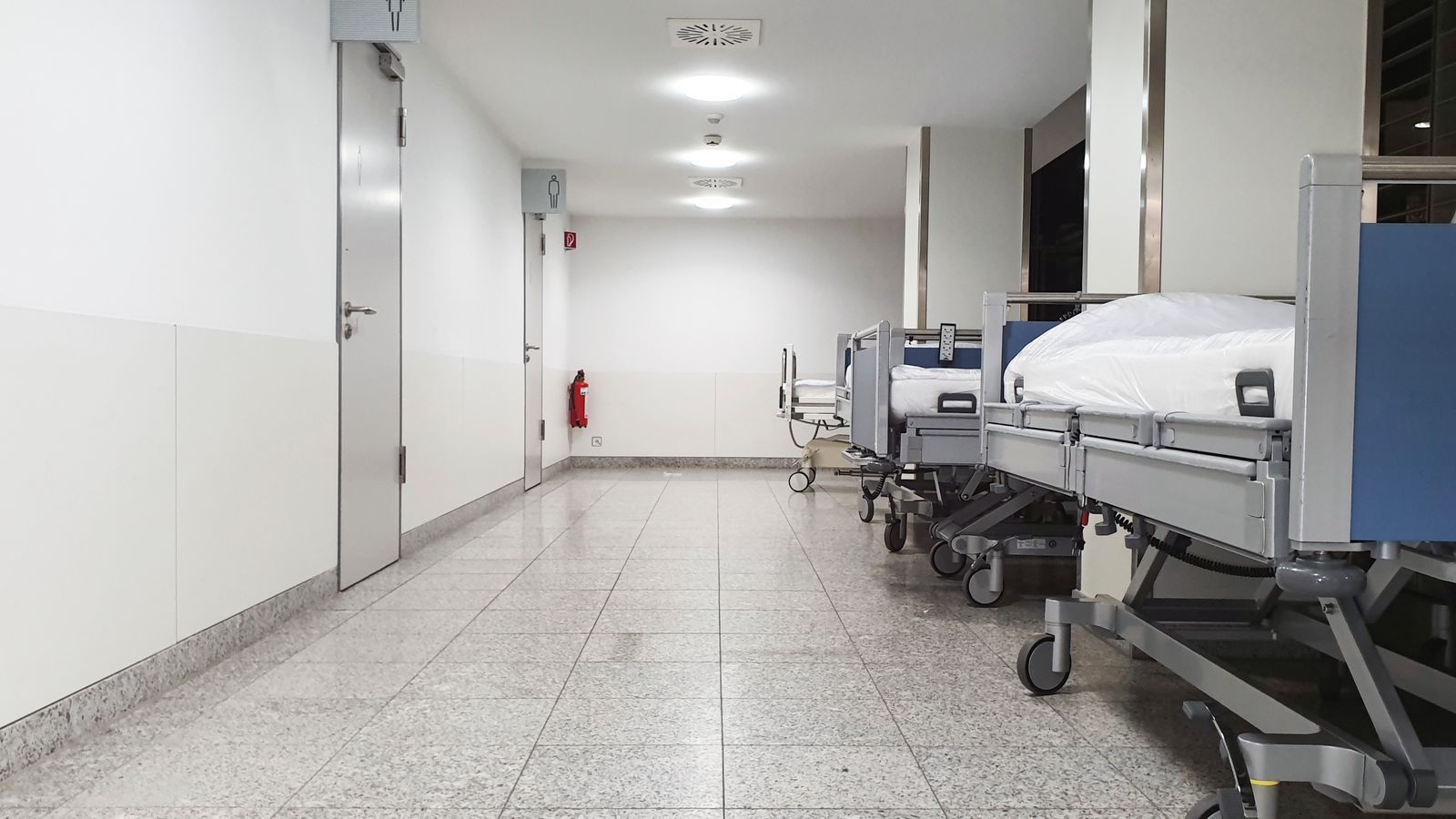 Empty hospital corridor with gurneys lined along the right wall and white walls under bright lights