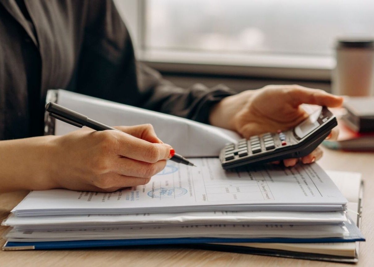 Hands writing on papers while using a calculator on a desk with a black pen and notebook stack