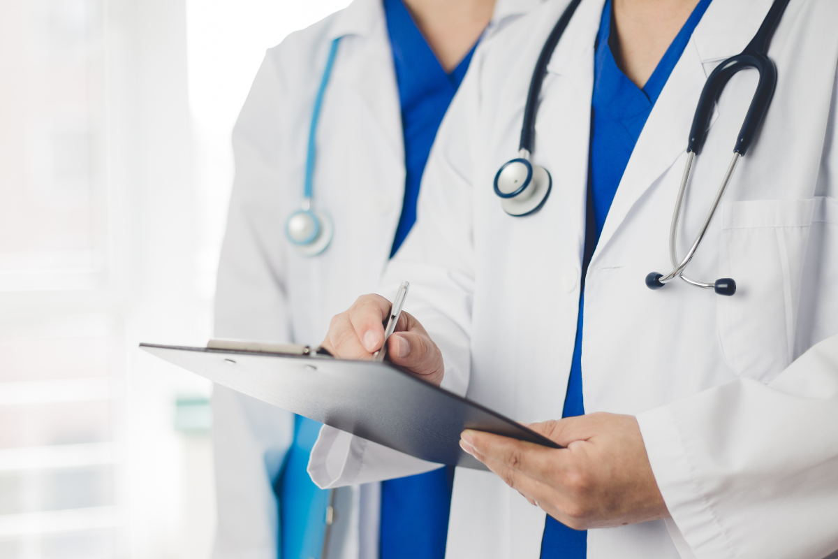Two doctors in white coats, holding a clipboard and stethoscope, indoors.