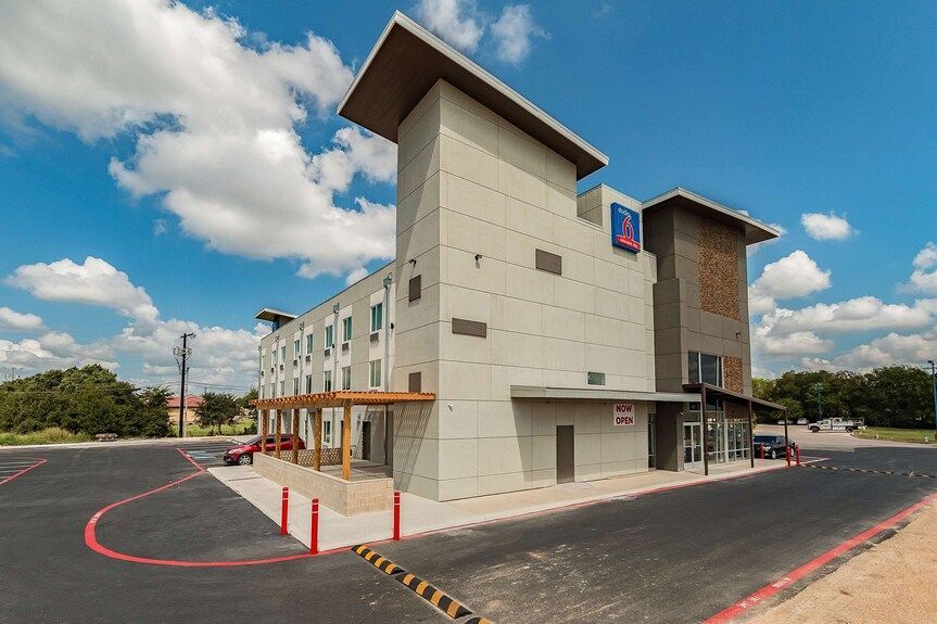 A modern three-story hotel with a beige concrete exterior, a tall architectural feature, and a paved parking lot.