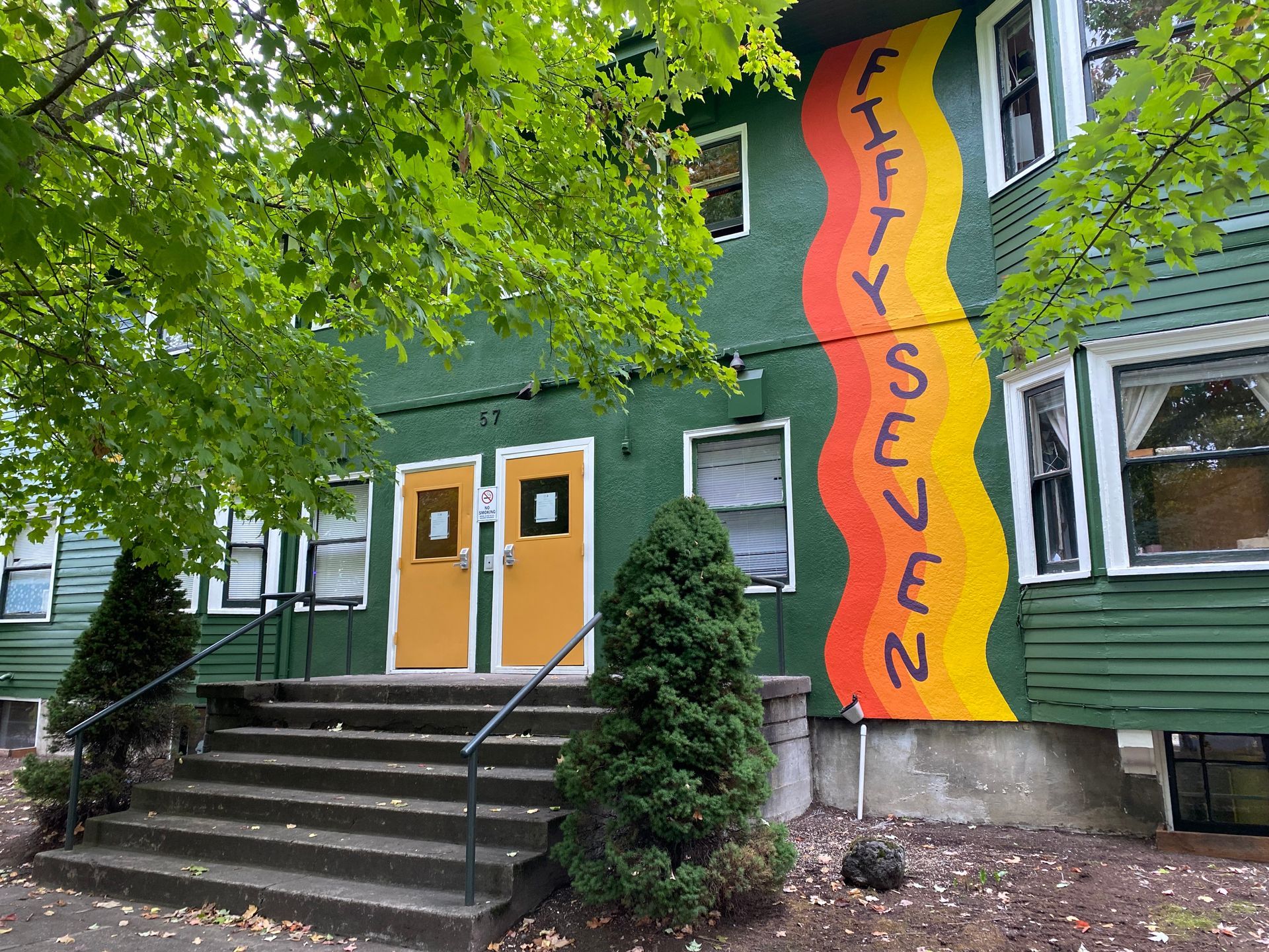 Colorful building entrance with orange doors, green siding, and a tall “Fifty Seven” mural beside steps and trees
