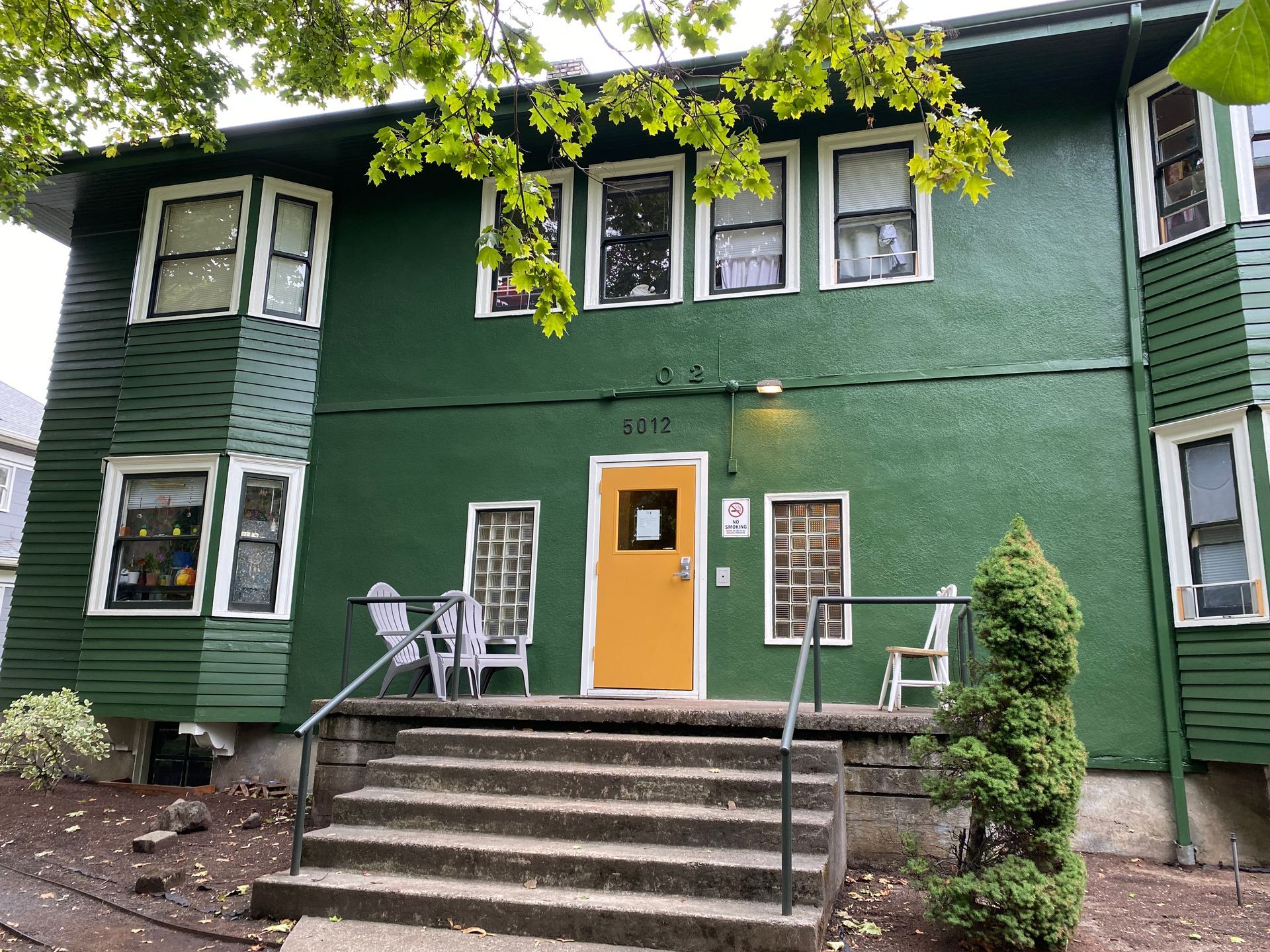 Green two-story apartment building with orange door, front steps, and small porch under leafy trees