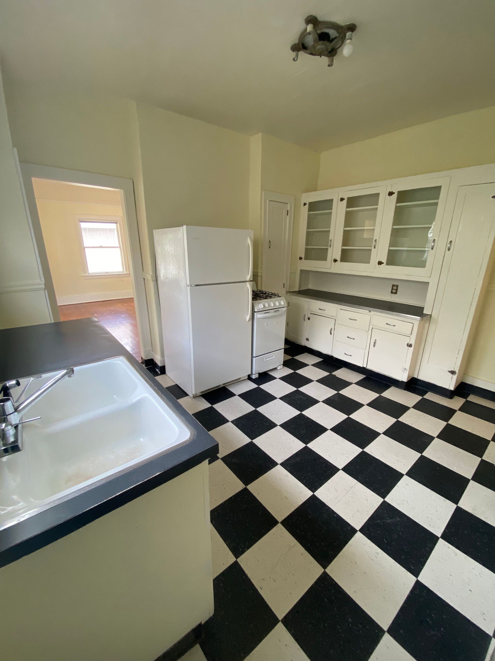Empty kitchen with black-and-white checkered floor, white cabinets, refrigerator, and sink counter.