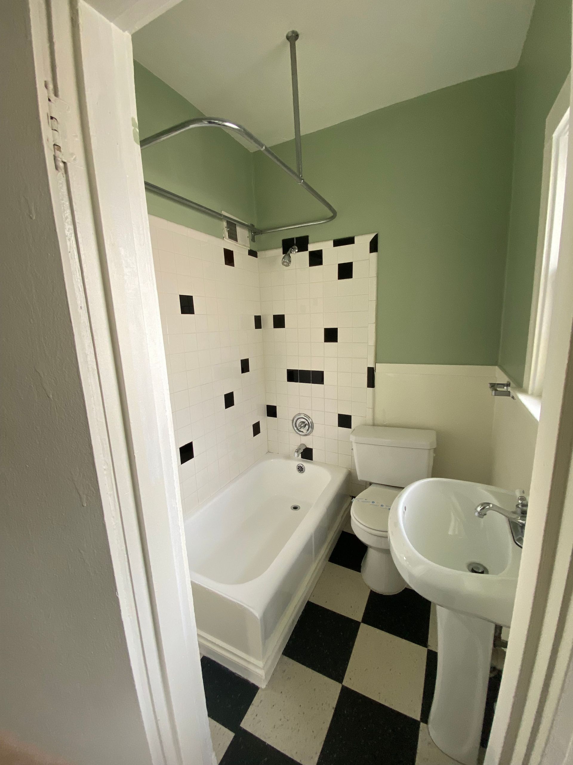 Small bathroom with white tub, pedestal sink, toilet, and black-and-white floor tiles.