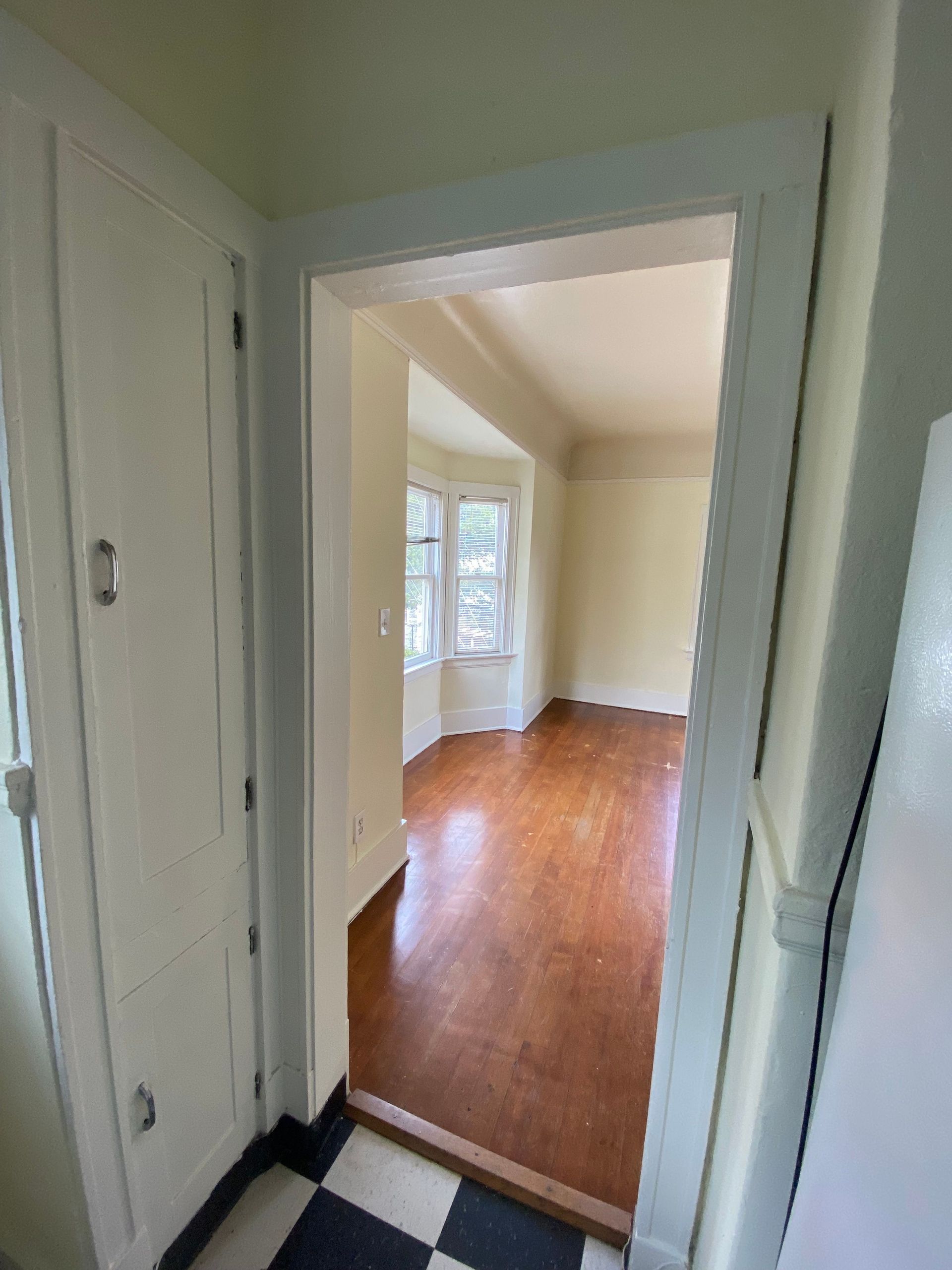 Narrow hallway opening into a bright empty room with hardwood floors and white trim.