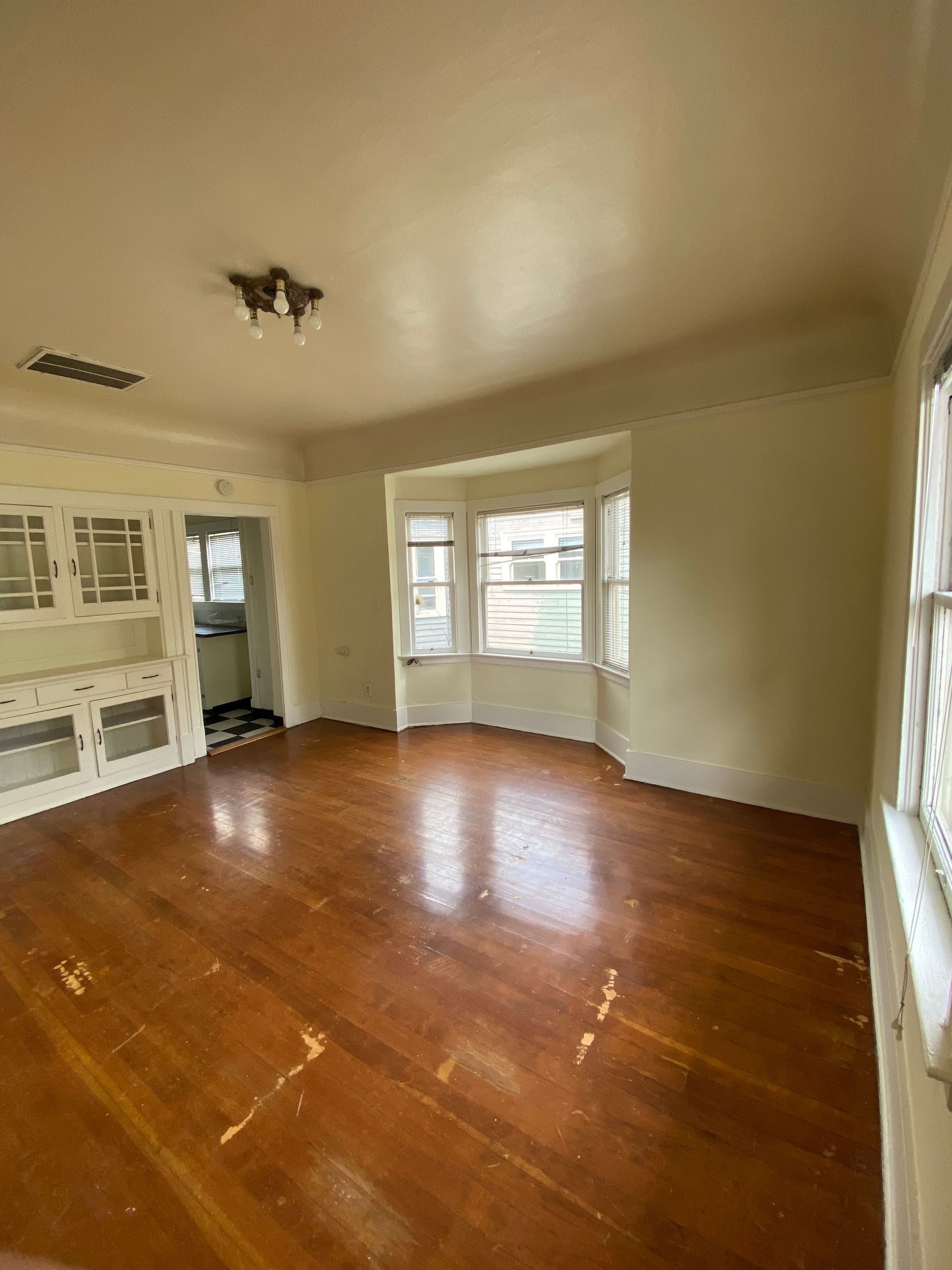 Empty living room with hardwood floors, cream walls, bay windows, and a ceiling light fixture