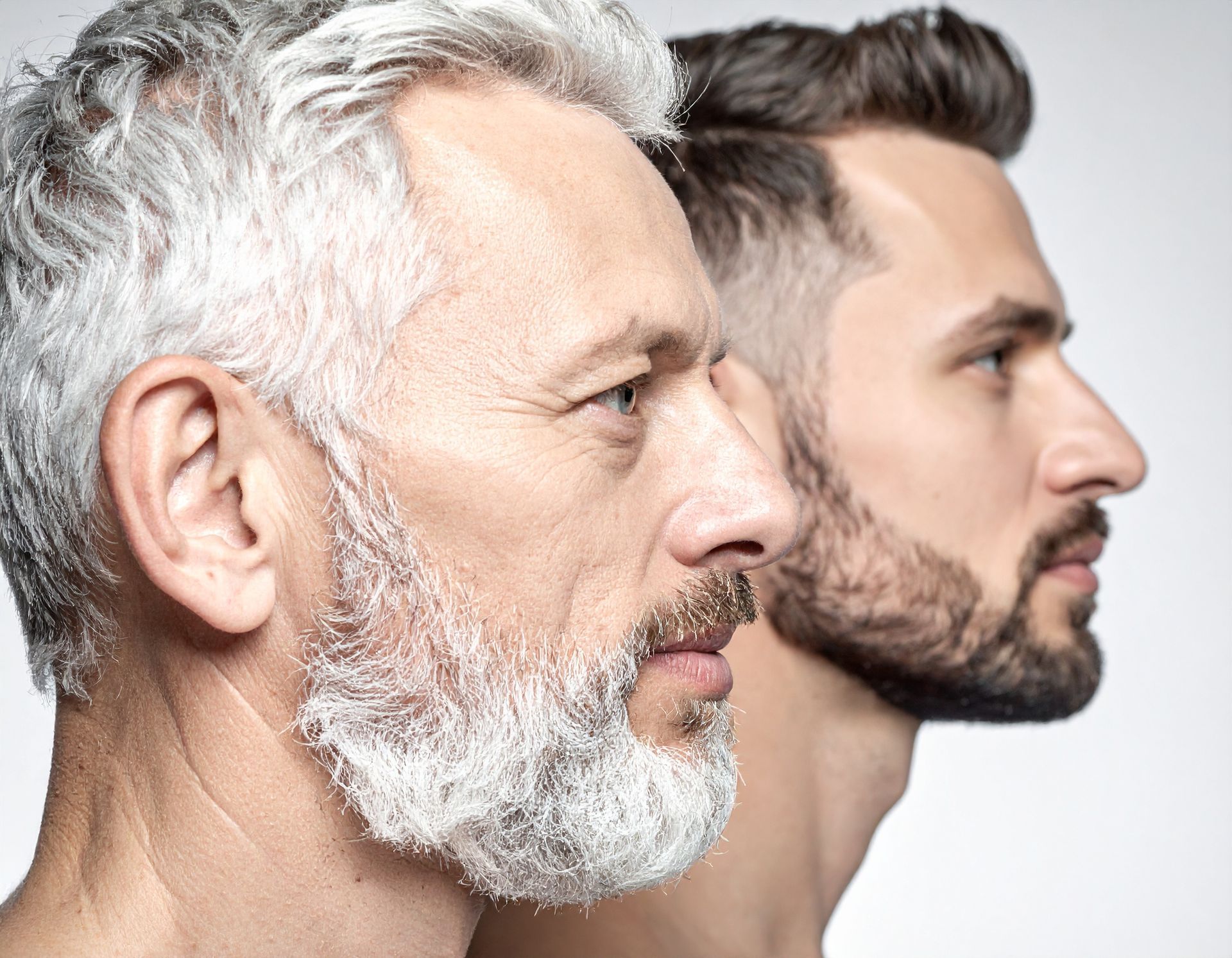 Two men, side profiles: older with gray hair/beard, younger with dark hair/beard, in front of white background.
