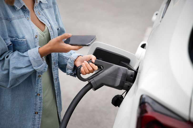 A woman is charging her electric car at a charging station while holding a cell phone.