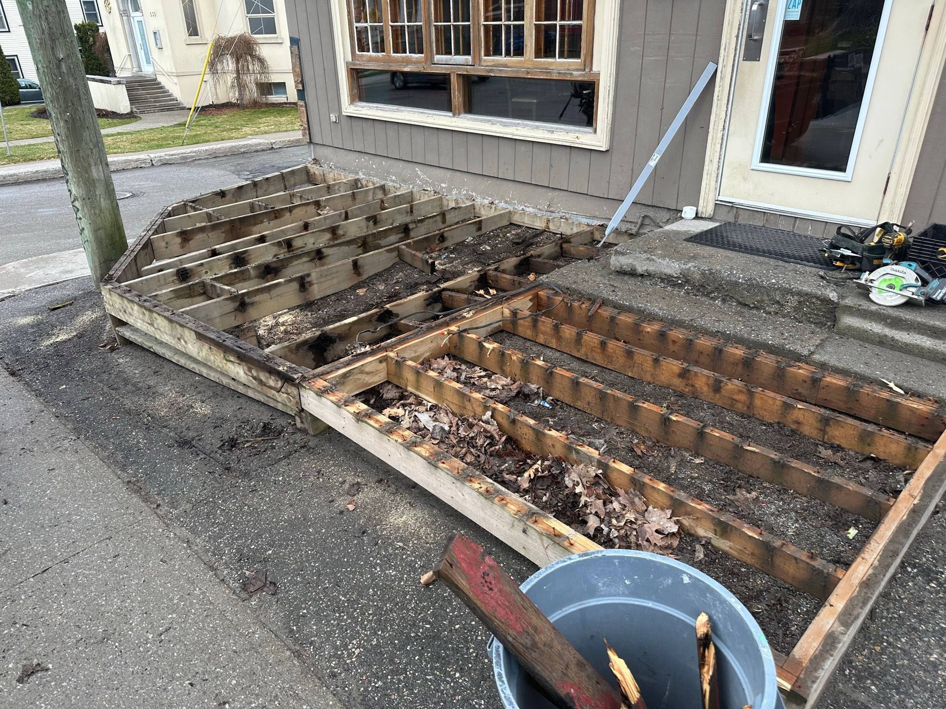 Porche et terrasse en bois démantelés devant un bâtiment ; débris et seau présents.