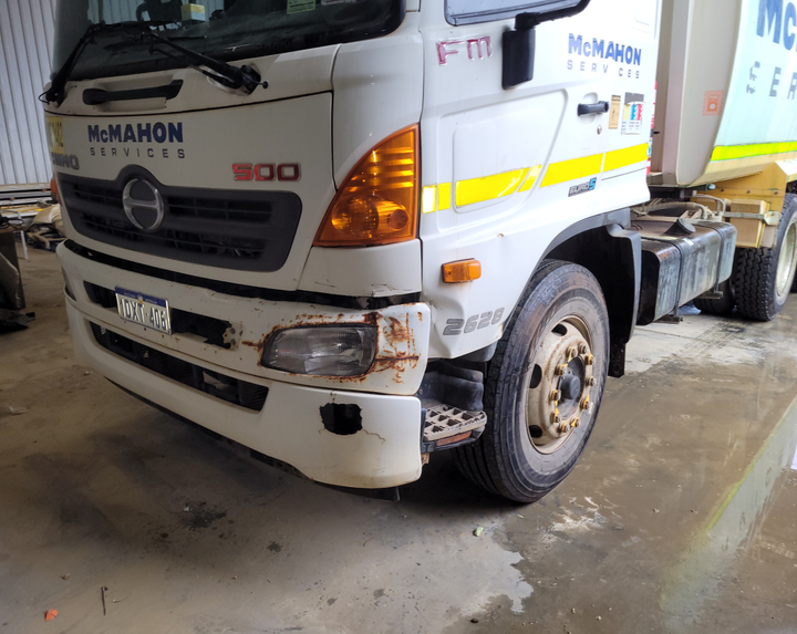A white truck with a broken bumper is parked in a garage.