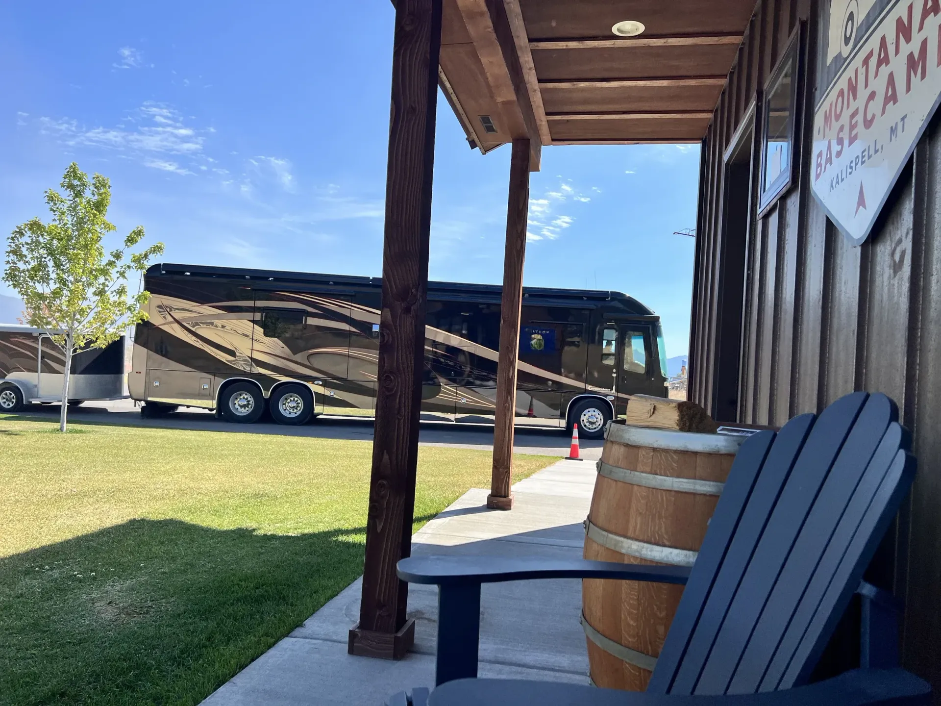Porch with wooden posts, a blue chair and barrel, overlooking a parked black tour bus on a sunny day