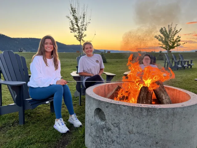 Three people sitting around a fire pit at sunset in a grassy outdoor area, with lounge chairs and hills in the background