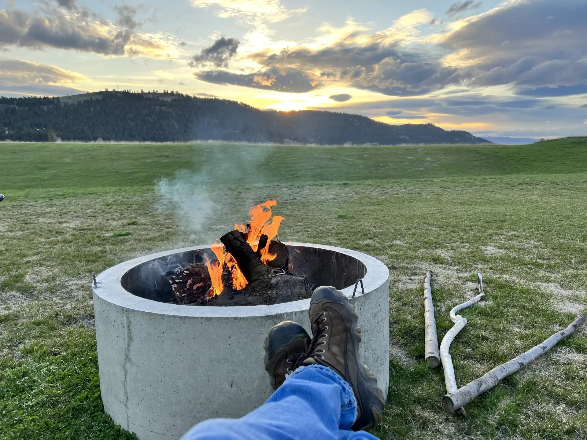 Fire burning in a concrete fire pit on a grassy field at sunset, with a booted foot in the foreground.