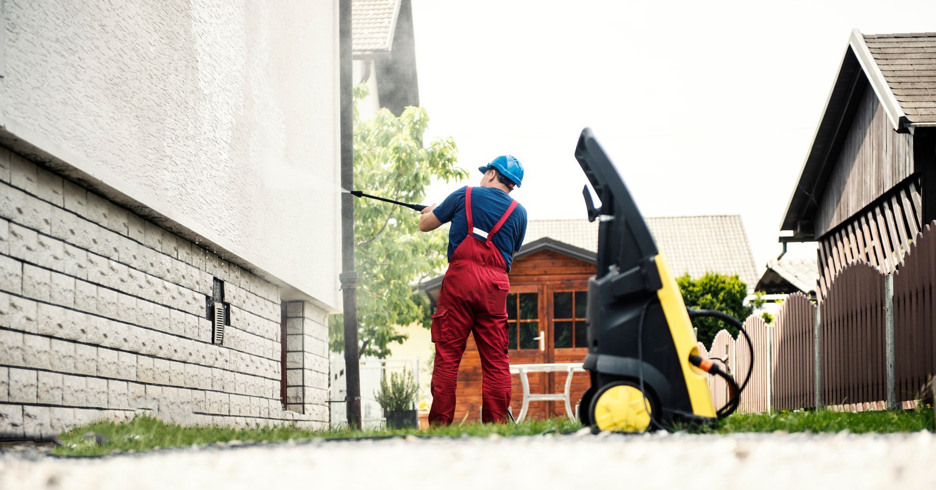 A man uses a pressure washer to clean the side of a house, spraying water against the wall.