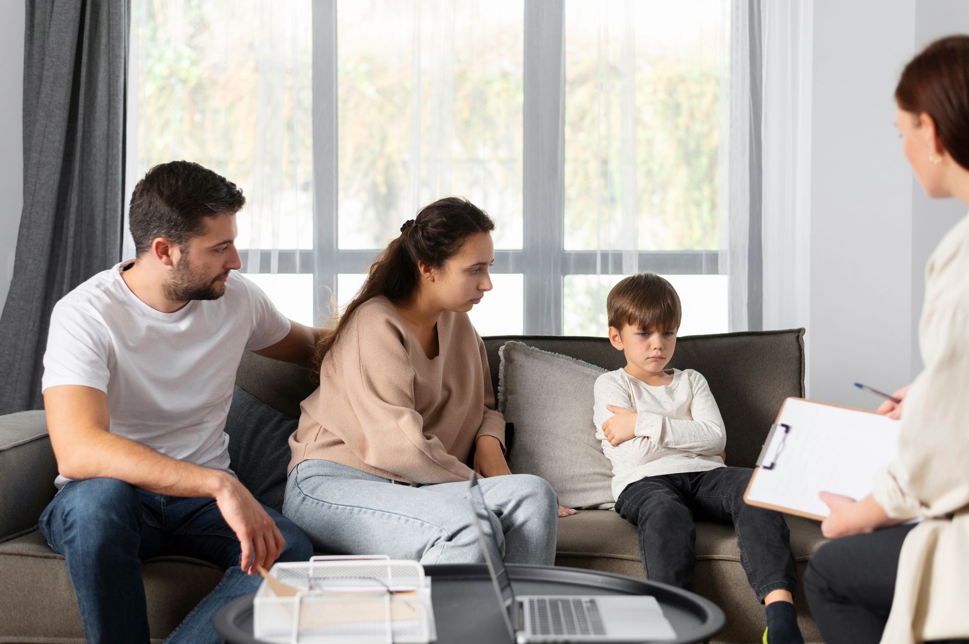Family, child, and therapist on a couch; the child looks upset, parents concerned, therapist takes notes.