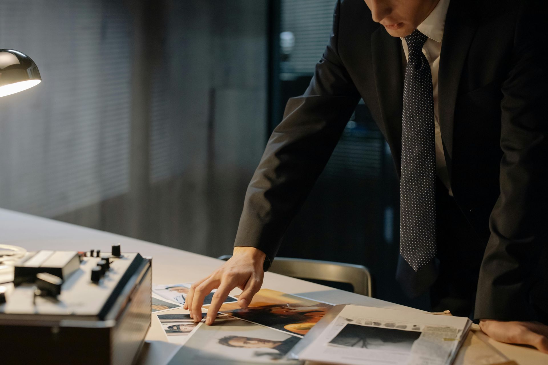 Person in suit examines photos on a desk in a dim office setting, next to a lamp and tape recorder.