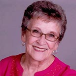An elderly woman wearing glasses and a pink shirt is smiling for the camera.