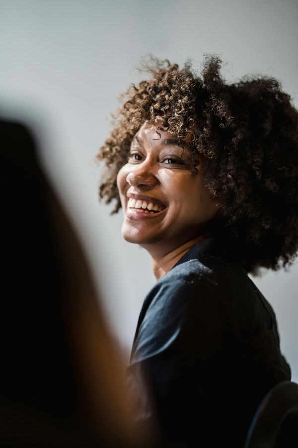 Smiling person with curly hair in a dark shirt, turned toward the camera against a soft gray background