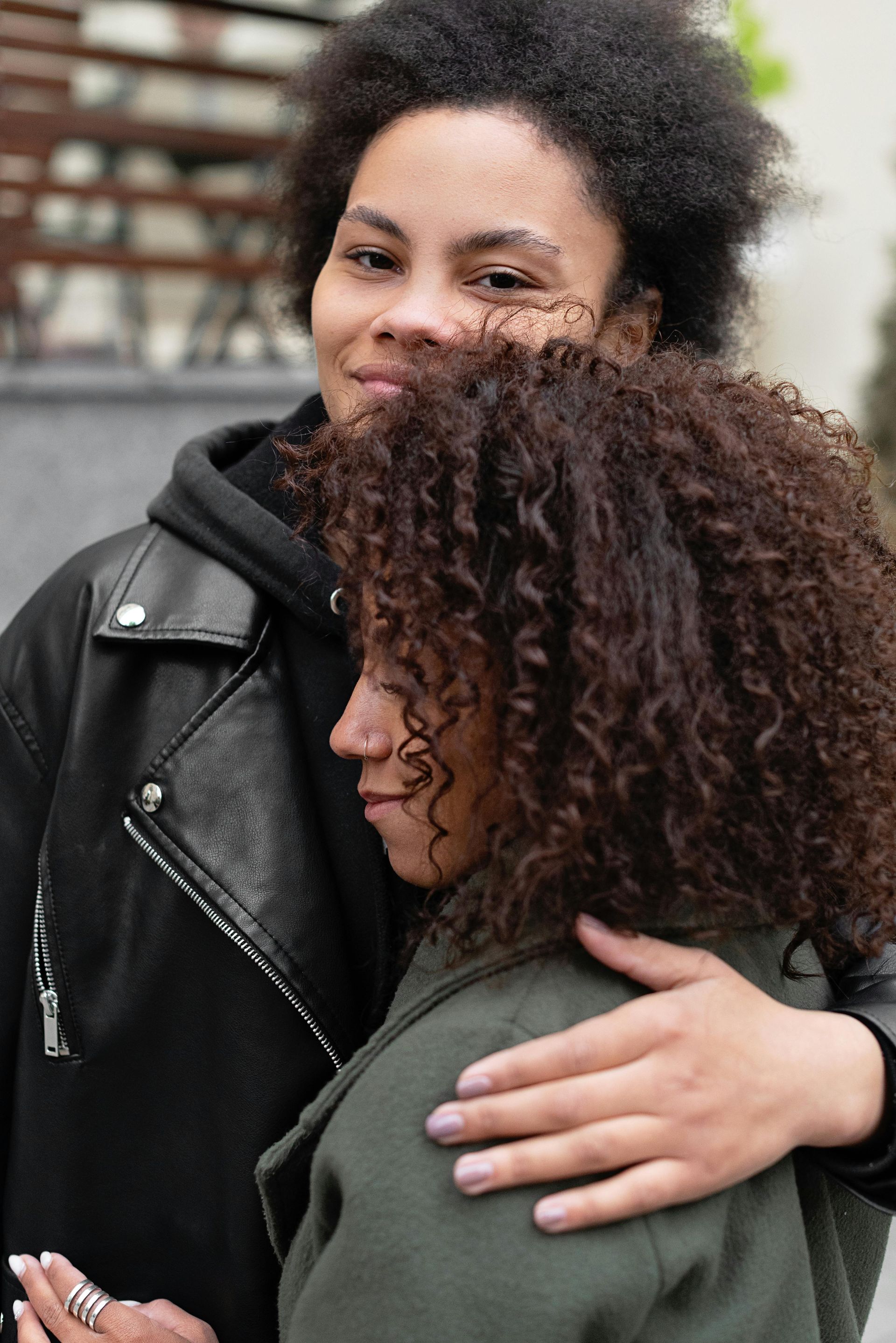 Two people hugging outdoors, one in a black leather jacket and the other with curly hair and a green coat.