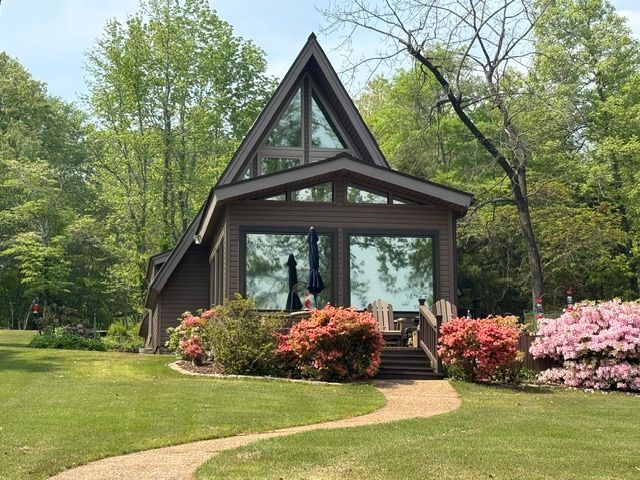 A small A-frame cabin with large windows, surrounded by green trees, lawn, and blooming pink shrubs.