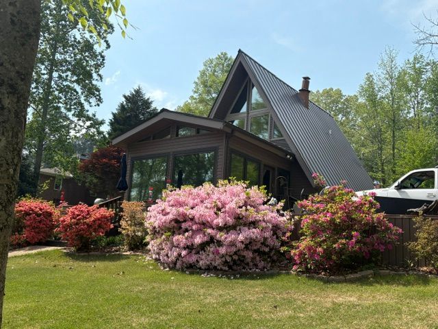 A cozy A-frame house with pink flowering shrubs and a green lawn under a clear blue sky.