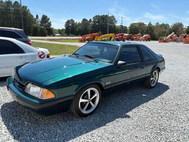 Dark green Ford Mustang coupe parked on gravel lot beside a white car under a sunny sky