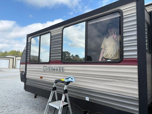 Silver and maroon camper trailer with tinted windows and a ladder parked on gravel under a blue sky